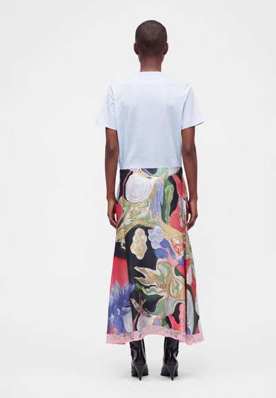 Personne debout de dos, portant un t-shirt blanc uni à manches courtes, une longue jupe à motif abstrait coloré et des bottes noires à talons.