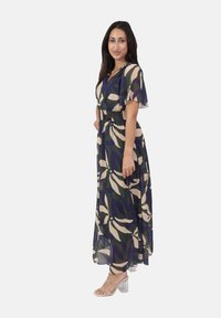 Kvinna i en lång, flödande klänning med stora krämfärgade och marinblå blommönster, som bär genomskinliga klacksandaler, stående i profil och leende.