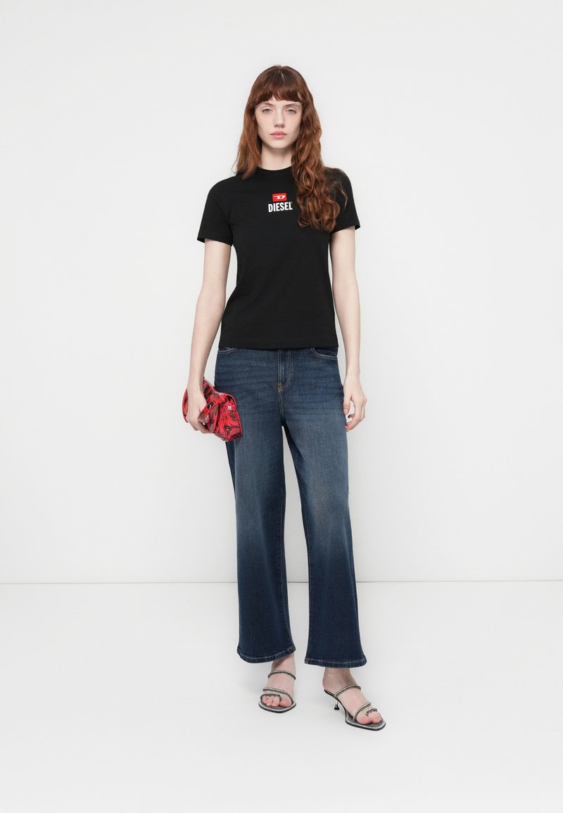 Donna che indossa una t-shirt nera Diesel, jeans a zampa blu, tiene una pochette rossa e indossa sandali neri con tacco e cinturini.