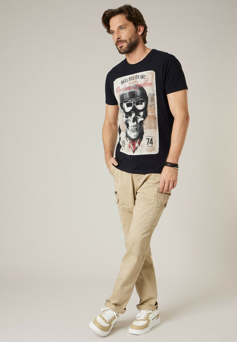 Homme portant un t-shirt noir avec un motif de crâne, un pantalon cargo beige et des baskets blanches, debout les mains dans les poches devant un fond neutre.