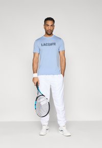 Camiseta azul claro con el logo "LACOSTE", pantalones deportivos blancos, zapatillas deportivas blancas, sosteniendo una raqueta de tenis con detalles en negro y azul.