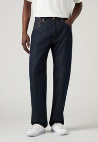 Mörkblå denimjeans med rak bendesign, prydda med orange sömmar, dragkedja och fem fickor. Bärs med vita sneakers.