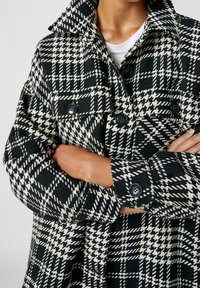 Manteau à carreaux noir et blanc avec un col, des poignets boutonnés et des poches. Matière texturée avec des motifs géométriques audacieux.