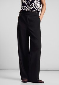 Svarta vida byxor med smidig tyg, elastisk midja och sidofickor, i kombination med en mönstrad topp och platta skor.