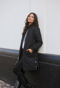 Black quilted long coat with a high collar and ribbed cuffs, featuring side pockets and snap buttons at the hem, worn with black trousers and boots.