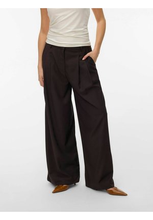 Kvinde iført sorte bukser med vide ben, hvid ærmeløs top og brune spidse sko, med den ene hånd i lommen.