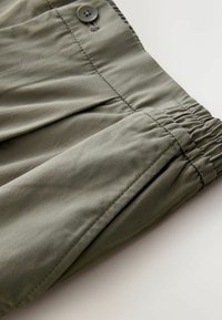 Green, lightweight fabric trousers feature an elastic waistband, button closure, and pleats for added shape and texture.