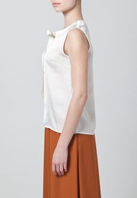 Krémová saténová bezrukávová blůza s mašlí na dekoltu, spojená s sukní v A-linii v odstínu rezavé. Hladká textury, minimalistický design.