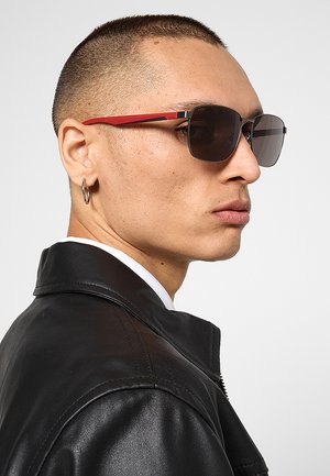 Uomo con capelli corti che indossa occhiali da sole quadrati con lenti scure e aste rosse, orecchino ad anello e giacca di pelle nera, rivolto verso destra.