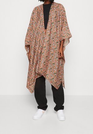 Personne portant un kimono ample à motif floral sur un haut noir et un pantalon, avec des baskets blanches, debout devant un fond clair uni.