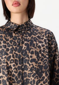 Leopard-print shirt in beige and black, featuring a classic collar, button closure, and a single chest pocket. Smooth fabric texture.