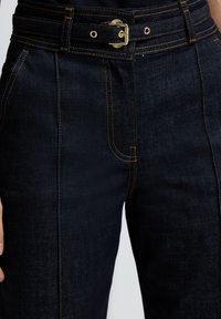 Tmavé džínové kalhoty s vysokým pasem, širokým páskem, kovovou sponou, viditelným švem a předními kapsami. Texturovaná tkanina, přizpůsobený tvar.