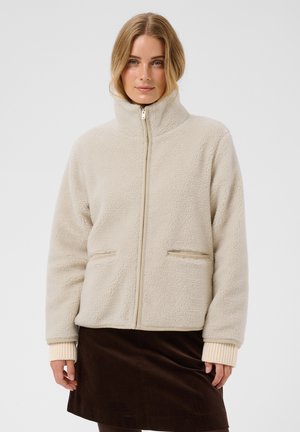 Femme portant une veste polaire beige à fermeture éclair avec un col montant et des poches avant, associée à une jupe marron foncé.