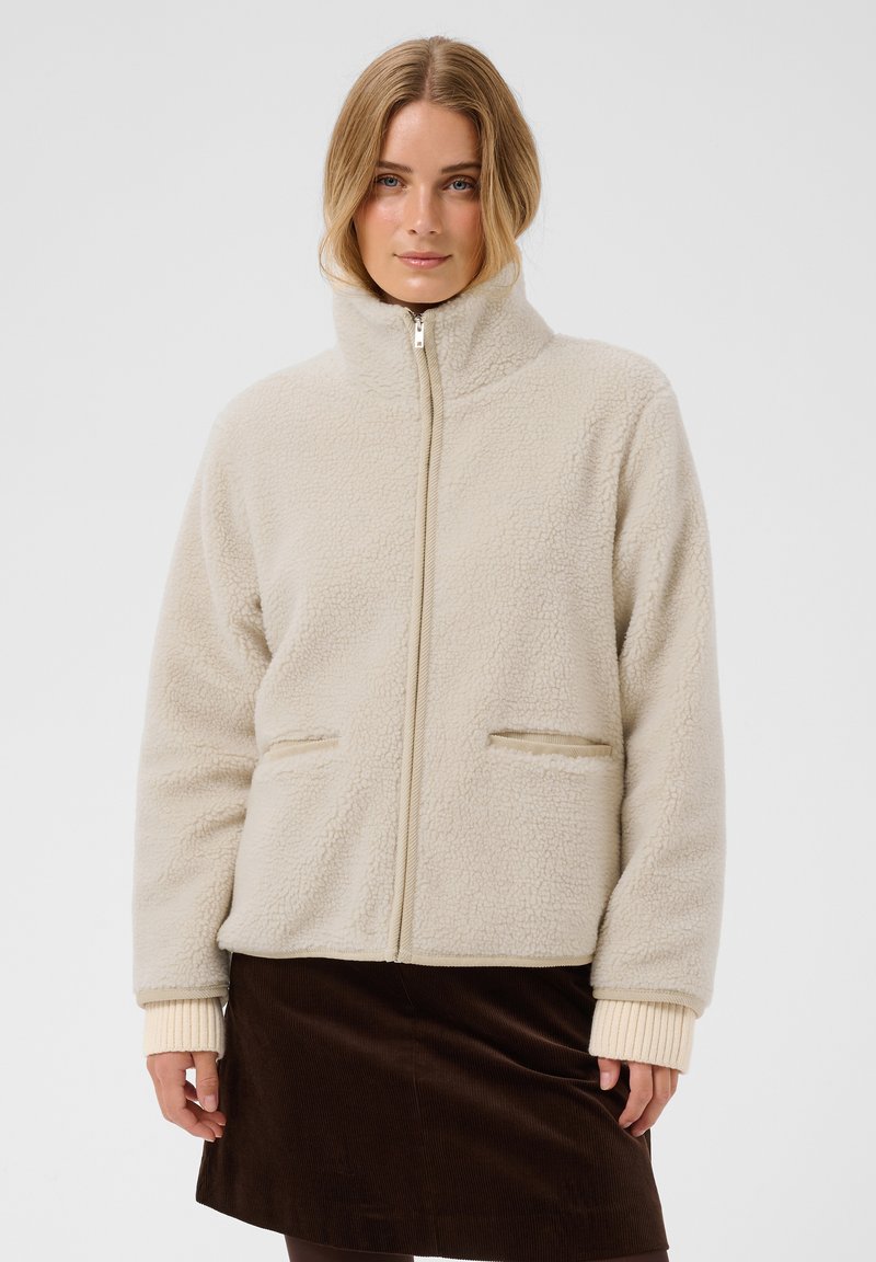 Femme portant une veste polaire beige à fermeture éclair avec un col montant et des poches avant, associée à une jupe marron foncé.