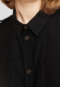 Schwarzes Hemd mit Knöpfen und Kragen, ausgestattet mit zwei vorderen Taschen, braunen Knöpfen und sichtbaren Nähdetails auf einem strukturierten Stoff.