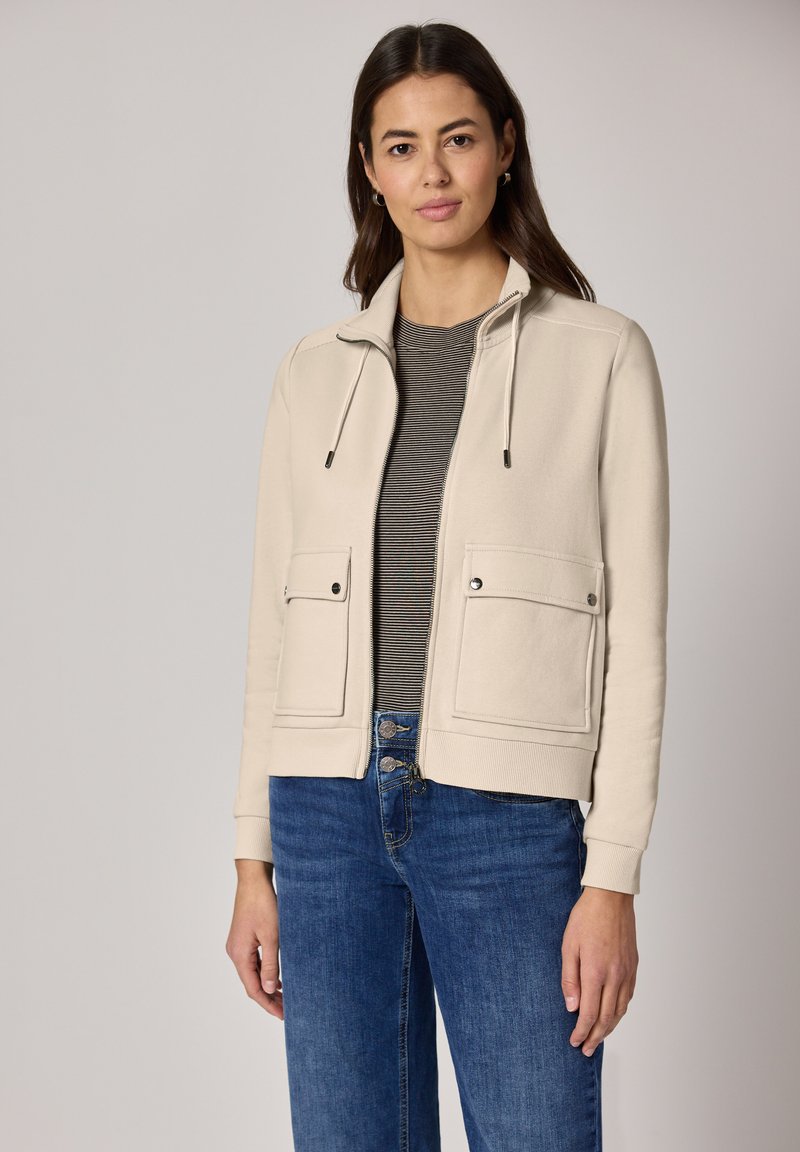 Veste bomber beige avec fermeture éclair à l'avant, capuche à cordon de serrage et deux grandes poches avant. Portée par-dessus une chemise grise rayée et un jean bleu.