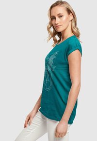 Tyrkysové bavlnené tričko s krátkymi rolovanými rukávmi a potlačou draka na prednej strane; skombinované s bielymi slim-fit nohavicami.