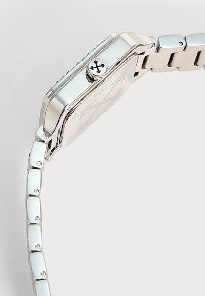 Bracelet et boîtier de montre en métal argenté avec une couronne latérale comportant un logo de flèche croisée sur un fond gris clair.