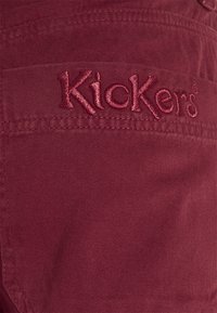 Nahaufnahme von kastanienbraunem Stoff mit gesticktem Markennamen "Kickers" in passendem Faden, die Textur und Nähte detailliert zeigend.
