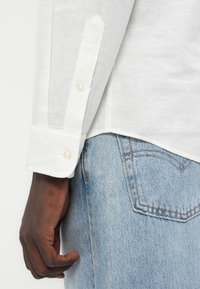 Λευκό πουκάμισο από λινό με μανσέτα με κουμπιά, εμφανιζόμενο δίπλα σε τζιν παντελόνι ανοιχτού μπλε χρώματος. Λεπτή υφή, εφαρμοστή γραμμή και διακριτική λάμψη.