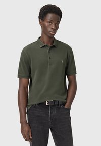 Mörkgrön polo-tröja med en texturerad yta, tre-knappsknäppning och diskret logotypdetalj på bröstet, i kombination med mörka jeans.
