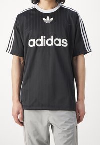 Musta urheilupaita, jossa valkoinen Adidas-logo, kolme valkoista raitaa hartioilla, pyöreä kaulus ja teksturoitu kangas. Yhdistetty vaaleanharmaisiin housuihin.