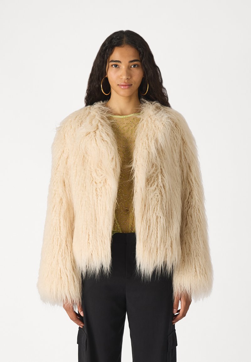Chaqueta de piel sintética en beige pálido con un diseño texturizado y desaliñado, que cuenta con un cuello de chal y mangas anchas, combinada con una blusa transparente con cuentas.