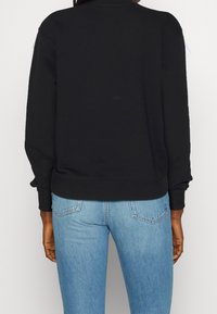 Sudadera negra con cuello y puños acanalados, que presenta un diseño liso. Combinada con jeans de mezclilla azul claros, mostrando una silueta ajustada.