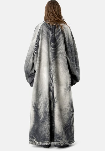 Person mit langen Haaren, der einen langen, übergroßen schwarz-weißen Acid-Wash-Denimmantel trägt, steht mit dem Rücken zu einer schlichten Hintergrundfläche.