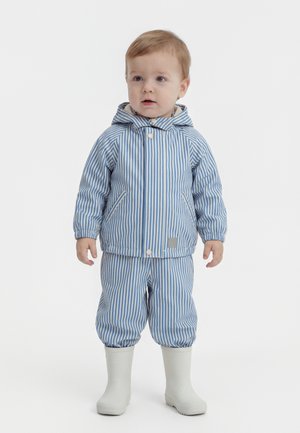 Småbarn stående iført blå og hvide lodrette stribede regnjakke og bukser med hvide gummistøvler mod en ensfarvet hvid baggrund.