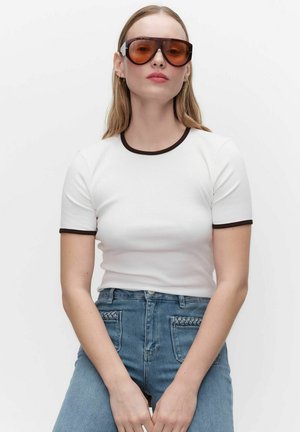 Frau mit langen blonden Haaren trägt große braune Sonnenbrille, weißes T-Shirt mit schwarzem Rand und hoch taillierte blaue Jeans mit geflochtenem Taschendetail.