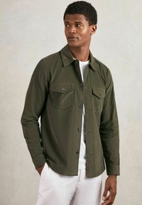 Olivgrüne Button-Up-Hemd aus strukturiertem Stoff, mit zwei Brusttaschen und einem klassischen Kragen, getragen über einem weißen T-Shirt.