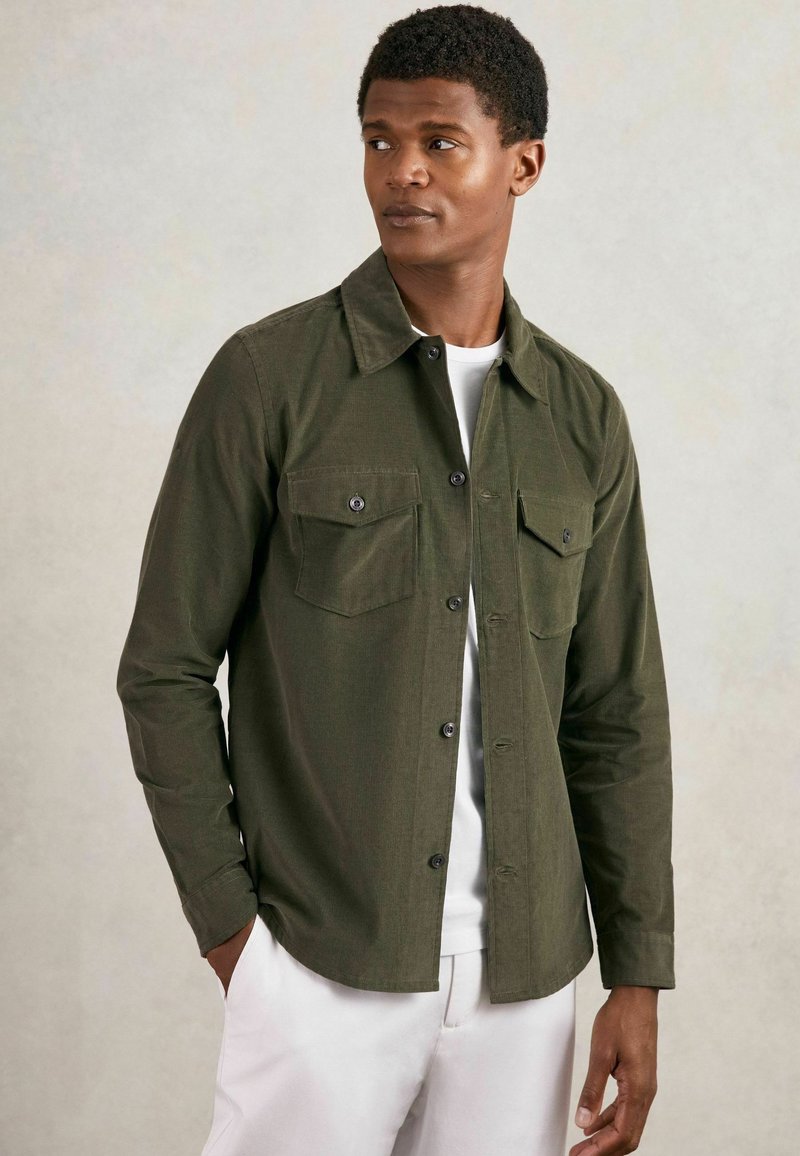 Olivgrüne Button-Up-Hemd aus strukturiertem Stoff, mit zwei Brusttaschen und einem klassischen Kragen, getragen über einem weißen T-Shirt.