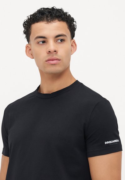 Junger Mann mit kurzen, lockigen Haaren, der ein schwarzes Dsquared2 T-Shirt mit kurzen Ärmeln trägt und nach links schaut, vor einem schlichten Hintergrund.
