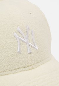 Cream-colored fleece cap with a structured shape, featuring a white embroidered "NY" logo on the front. Soft texture, minimal detailing.