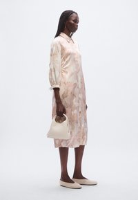 Φλοράλ φόρεμα σε απαλά ροζ και κρεμ αποχρώσεις, με γιακά και φουσκωτά μανίκια. Κρατάει μια μικρή λευκή τσάντα, φοράει ανοιχτό μπεζ παπούτσια.