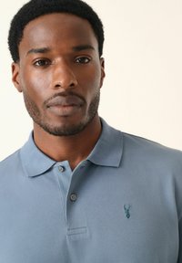 Polo en azul claro hecho de tejido texturizado, con un cuello, dos botones y un pequeño logo de ciervo bordado en el lado izquierdo del pecho.