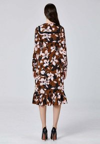 Robe fleurie brune à manches longues, avec un ourlet volanté, ornée de fleurs blanches et noires. Portée avec des chaussures à talons noirs.