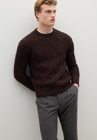 Pull marron en maille avec un motif torsadé, col rond et ourlet côtelé, porté avec un pantalon gris texturé. Le modèle se tient devant un mur neutre.