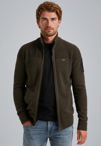 Maslinasto zeleni zip-up kardigan s teksturiranim pletenim materijalom, rebrastim manžetama i suptilnim oznakom na prsima, nosi se preko crne majice.