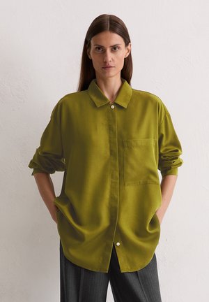 Groene oversized shirt met een puntige kraag, enkele borstzak en knopen. Gladde textuur, opgerolde mouwen en een relaxte fit.