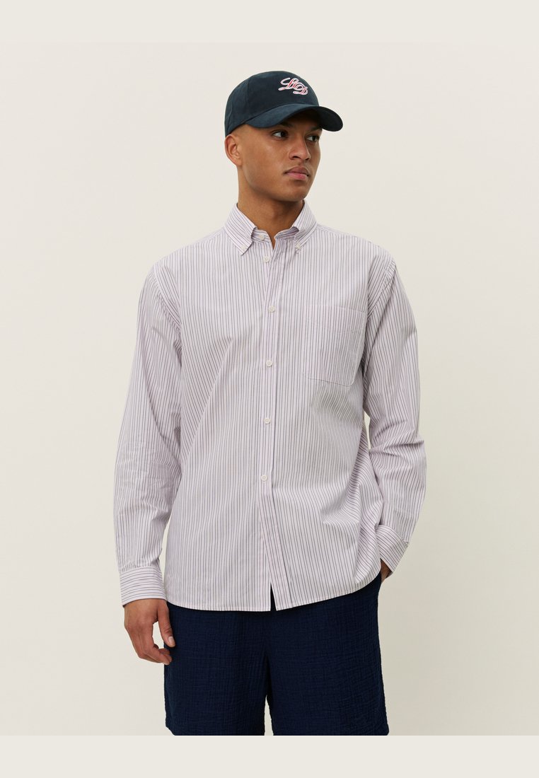 Camisa de botones a rayas en púrpura claro y blanco, con cuello puntiagudo, mangas largas y un bolsillo en el pecho. Llevada con pantalones cortos azul marino.