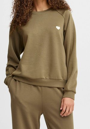 Kobieta ubrana w oliwkowo zieloną bluzę i pasujące spodnie, z prawą ręką w kieszeni i małym białym sercem na piersi bluzy.