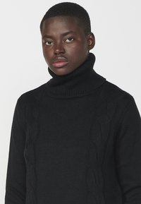Maglione a collo alto nero con un motivo a coste testurizzato e dettagli a treccia sui lati. Realizzato in un materiale morbido e spesso.
