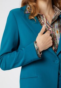 Blazer turquoise avec une fermeture à un seul bouton et deux poches avant, associé à une chemise à motifs multicolores avec un col contrastant.