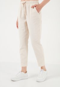Light beige, lightweight fabric pants with an elastic waistband, side pockets, and a slightly cropped, tapered fit. Finished with frayed hems.