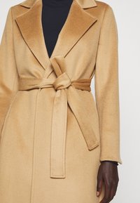 Femme portant un manteau en laine ceinturé couleur camel par-dessus un haut noir avec une main visible à son côté.