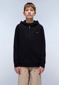 Sweat à fermeture éclair noir avec une poche avant, des poignets côtelés et un petit emblème décoratif sur la poitrine gauche. Porté avec un pantalon beige ample.
