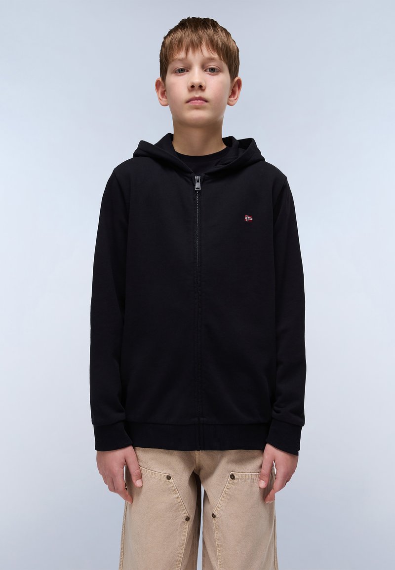 Sweat à fermeture éclair noir avec une poche avant, des poignets côtelés et un petit emblème décoratif sur la poitrine gauche. Porté avec un pantalon beige ample.