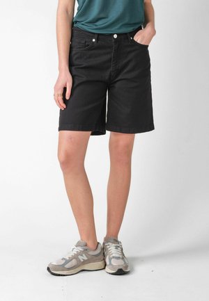 Person iført sorte shorts til knæet, turkis trøje, grå og hvide sneakers, stående med den ene hånd i lommen mod en ensfarvet baggrund.